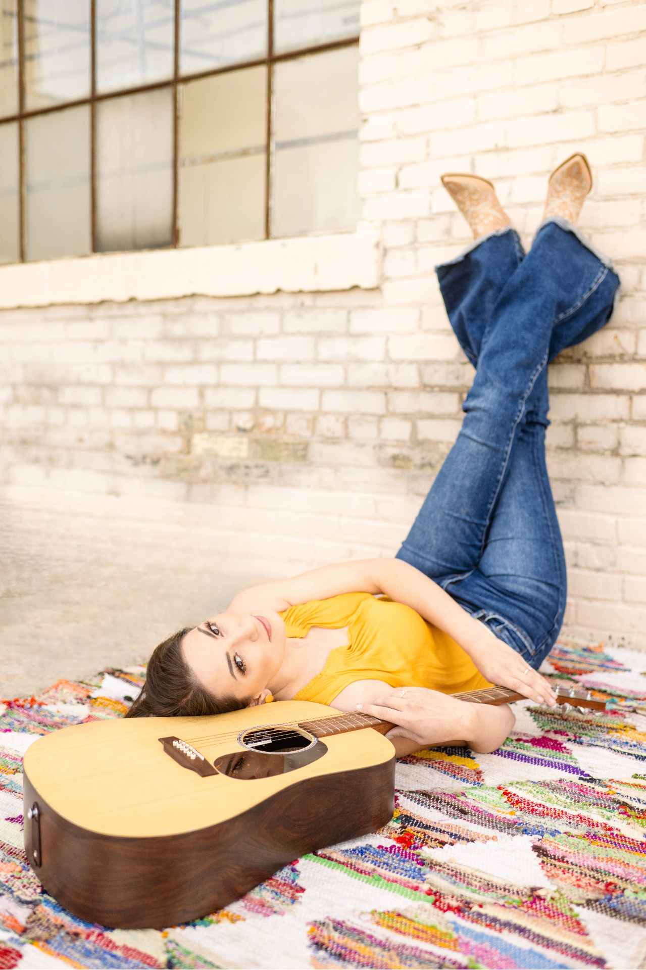 Anna Marie with guitar on a colorful rug, brick wall